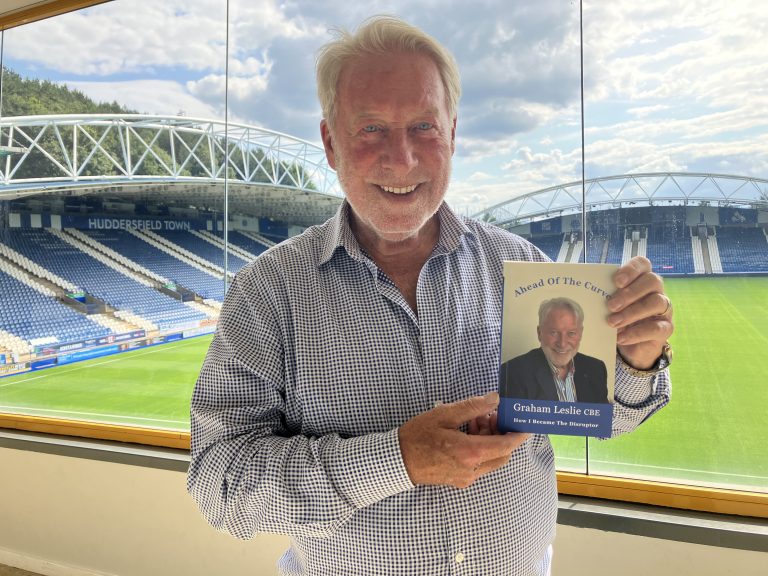 Graham Leslie holding a copy of his autobiography Ahead Of The Curve at the stadium he had the vision to create in the early 1990s